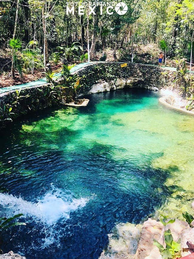 Vista vertical de un cenote abierto con agua azul y verde brillante, mostrando un chorro de agua cayendo en primer plano y un muro de piedra al fondo.