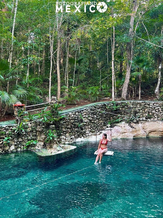Fotografía tomada desde la orilla que muestra a una mujer sentada en un columpio de madera suspendido sobre las aguas azules de un cenote abierto, rodeada por un muro de piedra y selva.