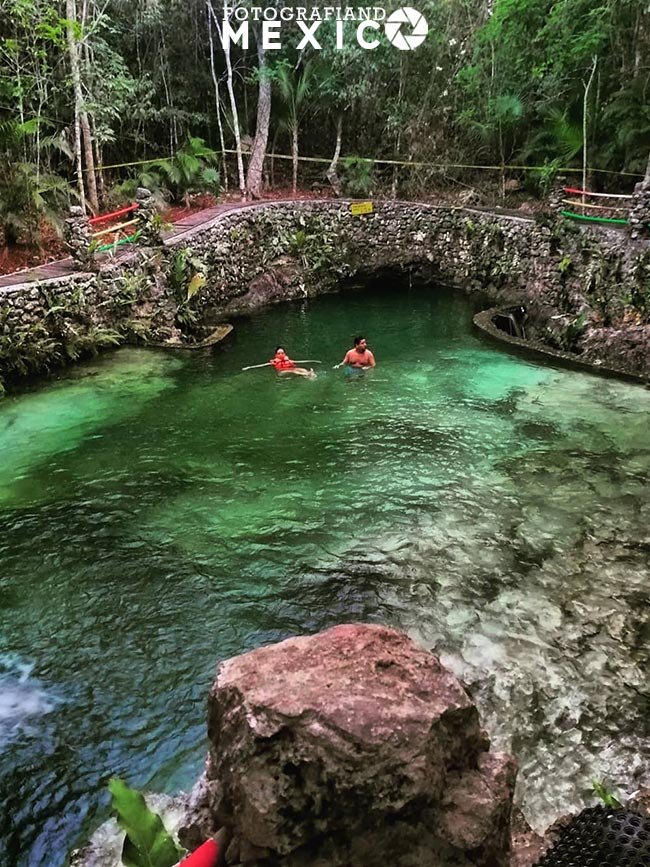 Vista en picada desde una roca que muestra a una pareja nadando en un cenote de agua verde transparente, rodeada por un muro curvo de piedra y árboles densos.