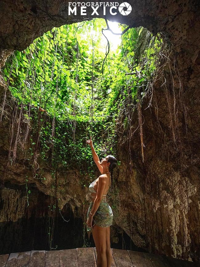 Vista desde el interior de una caverna que muestra a una mujer de perfil sobre una plataforma de madera, señalando hacia un tragaluz circular en el techo cubierto de enredaderas y raíces colgantes.