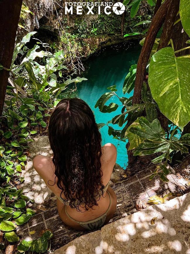 Vista desde arriba de una mujer de espaldas sentada en escalones de piedra caliza mirando hacia una poza de agua turquesa rodeada de plantas verdes silvestres.