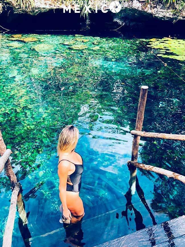 Mujer en traje de baño negro bajando escaleras de madera hacia el agua verde transparente de un cenote.