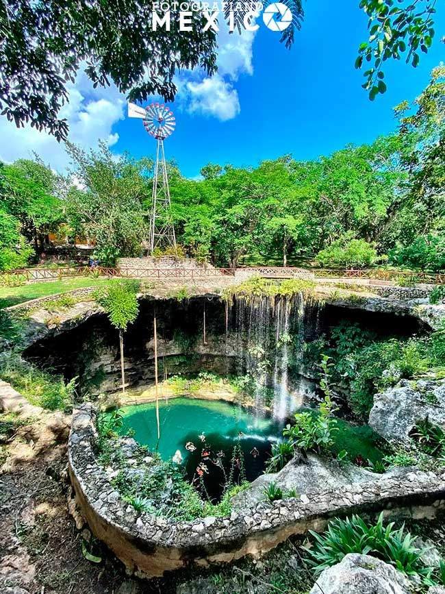 Cenote circular con una cascada y un molino de viento alto al fondo rodeado de selva.