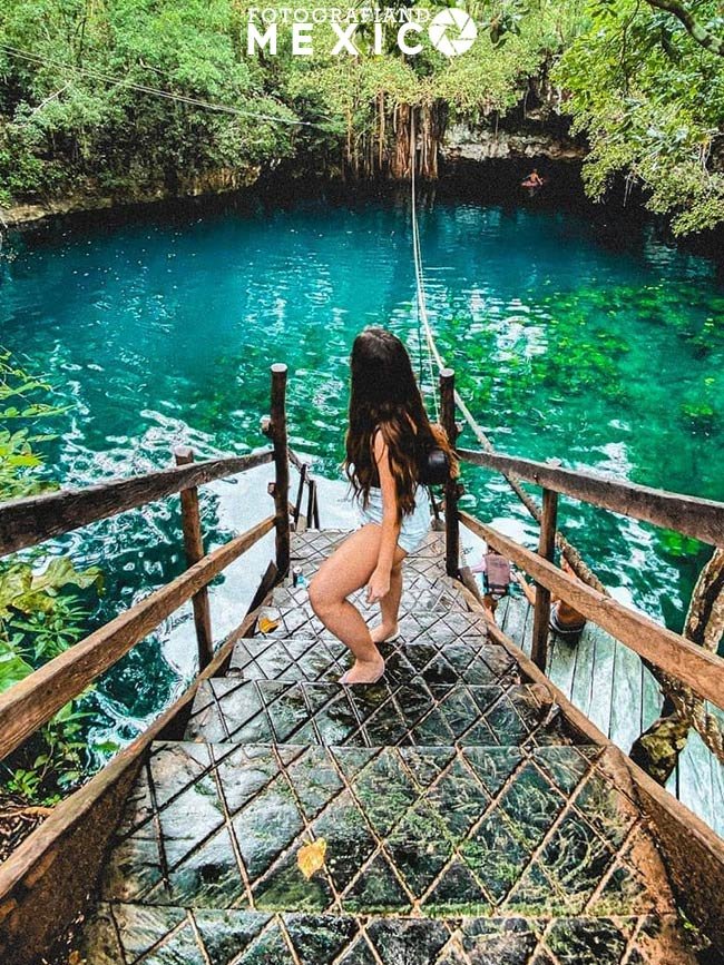 Vista desde arriba de una mujer bajando escalones de piedra caliza con pasamanos de madera hacia un cenote de agua azul y verde transparente rodeado de selva.
