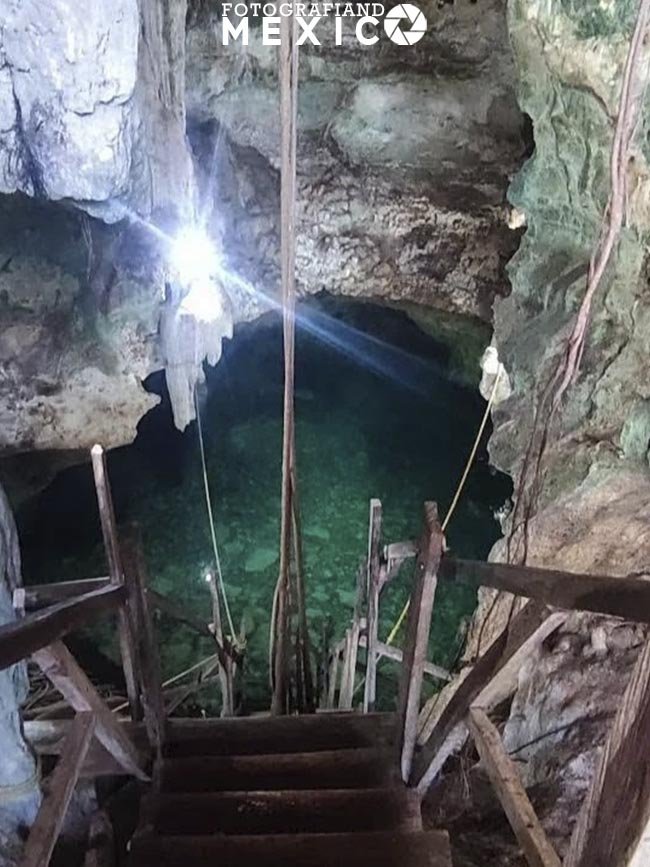 Escaleras rústicas de madera que descienden hacia la poza de agua verde cristalina iluminada artificialmente dentro del Cenote X'tojil.
