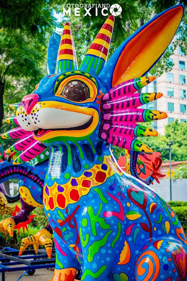 Escultura brillante de un alebrije con cabeza de conejo azul y cuernos decorado con patrones de flores coloridas en una plaza