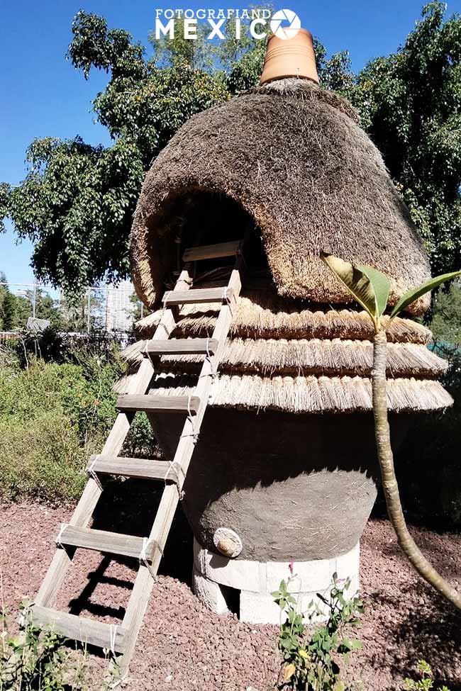Estructura tradicional de un cuexcomatl o granero azteca de barro con techo de paja y una escalera de madera apoyada en un jardín.