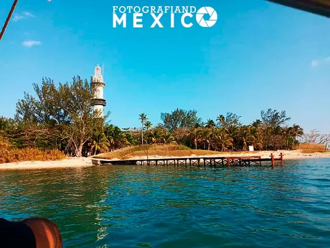 Vista desde una lancha que muestra un muelle rústico de madera sobre el mar turquesa, conectado a una playa de arena blanca con palmeras y un faro alto bajo un cielo azul despejado.
