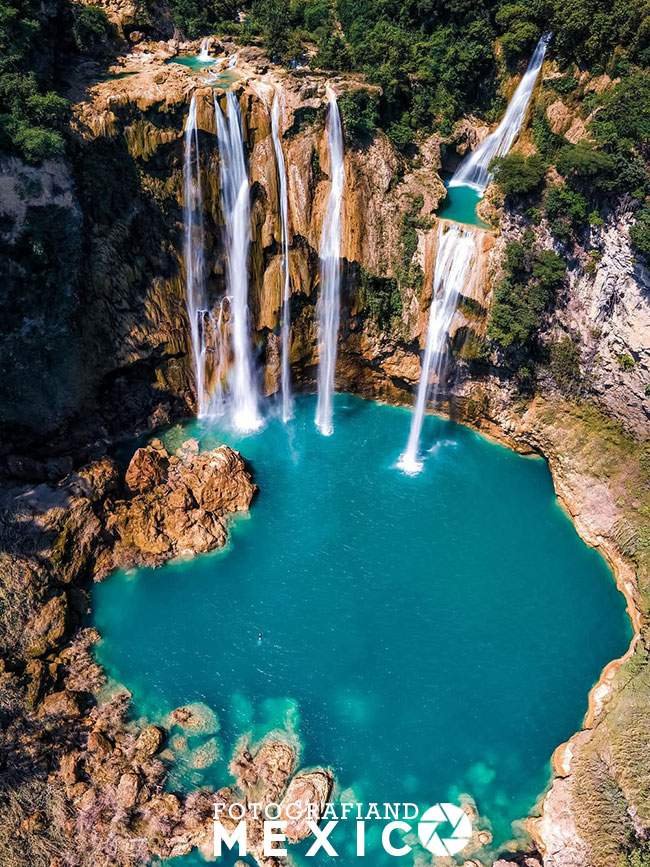Vista aérea vertical de una enorme poza circular de agua azul turquesa cristalina alimentada por múltiples cascadas altas que caen de acantilados rocosos rodeados de selva.