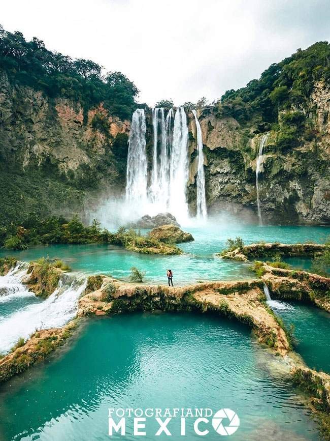 Vista frontal de un viajero parado sobre un dique natural de roca caliza rodeado de múltiples albercas escalonadas de agua turquesa frente a una colosal cascada de treinta metros.