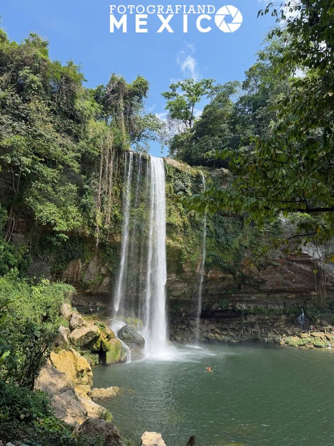 Vista frontal de una alta cascada que se desploma verticalmente desde un acantilado cubierto de vegetación hacia una poza de agua verde rodeada de densa selva y rocas.
