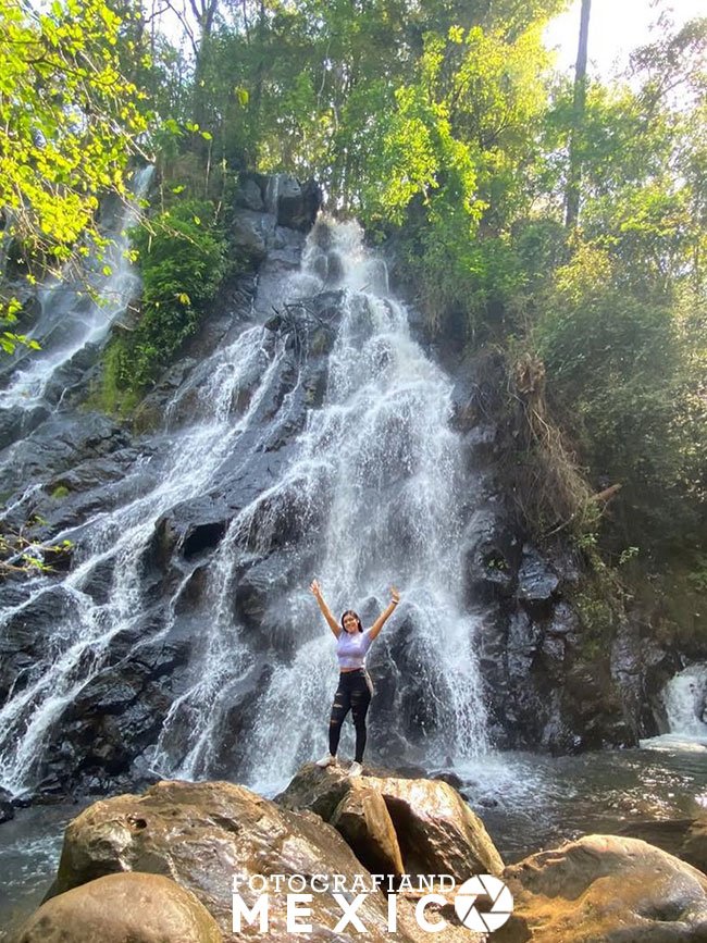 Una mujer con los brazos levantados posa sobre grandes rocas frente a una colosal cascada que se desliza por una muralla de piedra negra rodeada de densos árboles verdes.