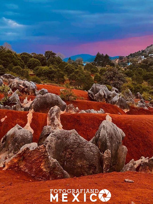 Perspectiva vertical de formaciones de piedra caliza gris esparcidas sobre lomas de arena roja bajo un cielo tormentoso con un resplandor rosado en el horizonte.