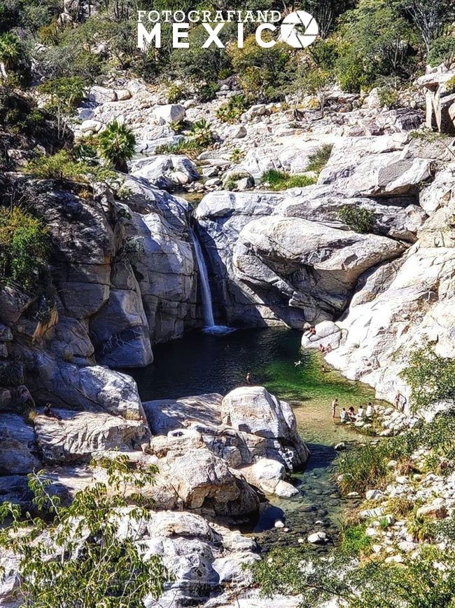 Vista panorámica de una cascada que cae entre colosales formaciones de roca blanca hacia una poza natural verde cristalina rodeada de vegetación desértica.