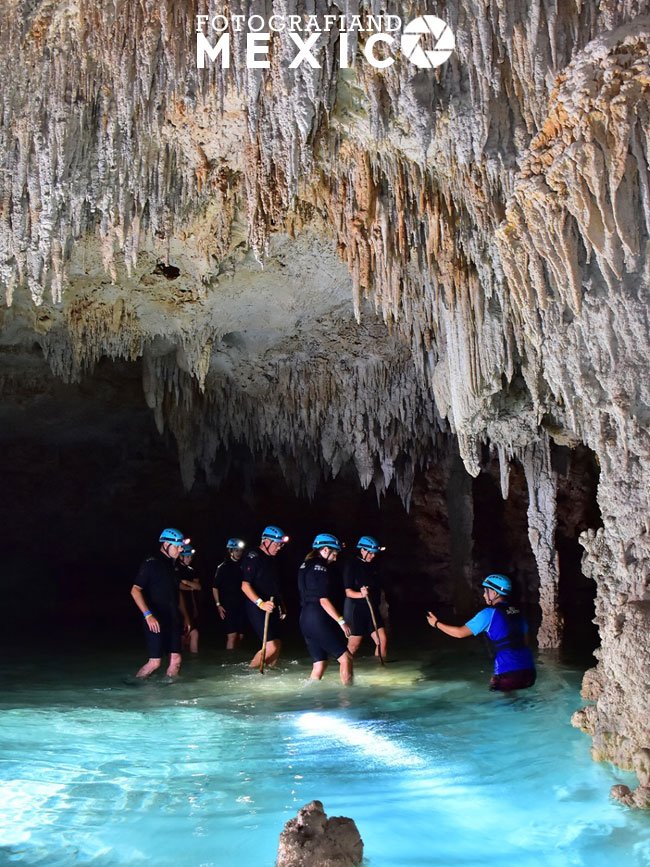 Un grupo de excursionistas con trajes de neopreno negros y cascos azules camina con el agua a la cintura dentro de una caverna inundada rodeada de estalactitas.