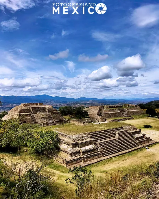 Vista panorámica de Monte Albán, que es una antigua ciudad construida por los zapotecas en la cima de una montaña, donde se pueden apreciar las pirámides, los palacios, los templos, los juegos de pelota, los altares, las tumbas y los petroglifos, bajo un cielo azul.