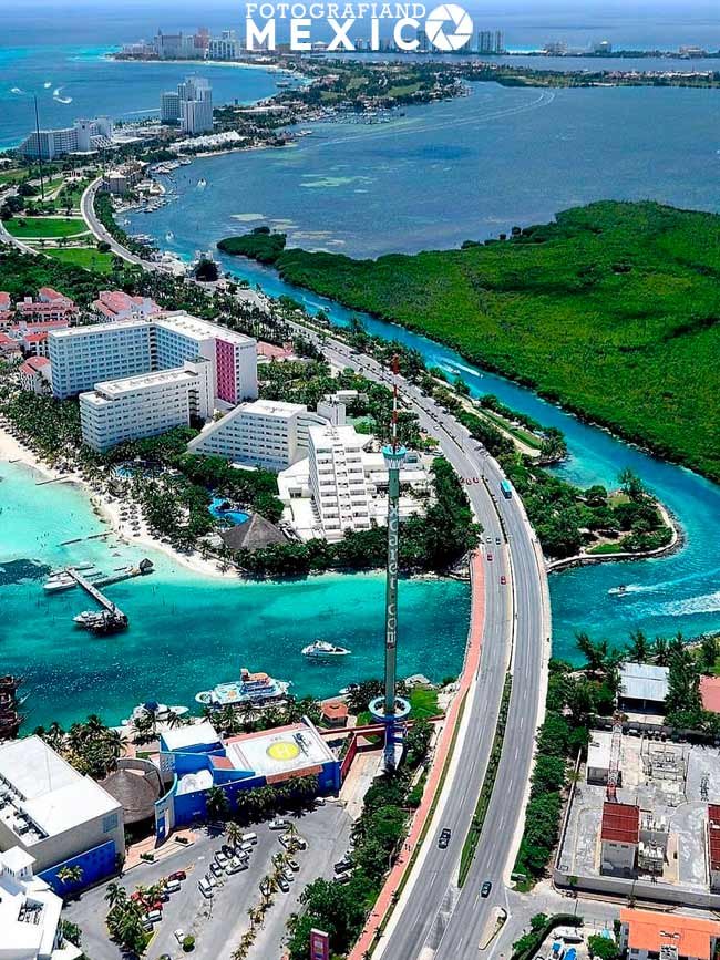 Vista aérea del inicio del Boulevard Kukulcán en Cancún cruzando el puente sobre el canal, con la Torre Escénica a un costado, hoteles de playa y la laguna Nichupté.