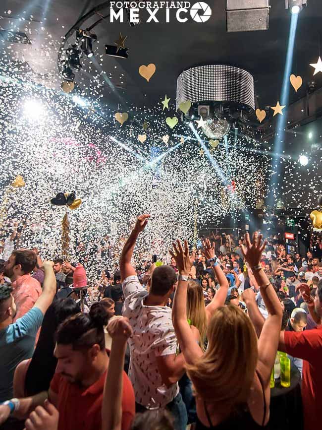 Una multitud de jóvenes de espaldas bailando con las manos en alto dentro de un club nocturno lleno de luces, bajo una lluvia masiva de confeti blanco y decoraciones de corazones colgantes.