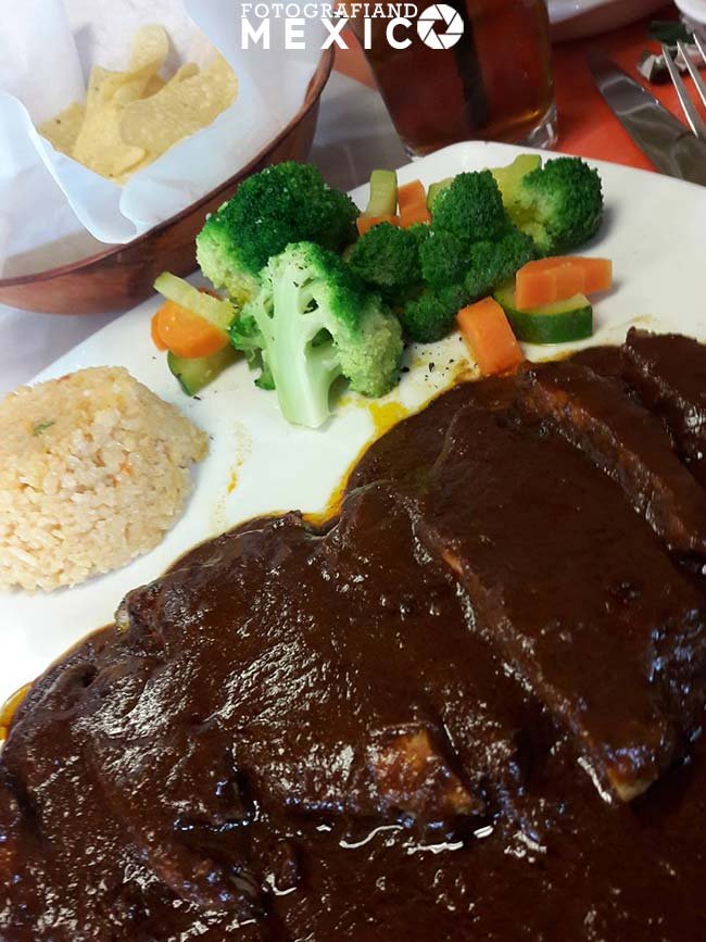 Pollo bañado en mole oscuro espeso con guarnicion de arroz y verduras al vapor servido en un plato blanco.