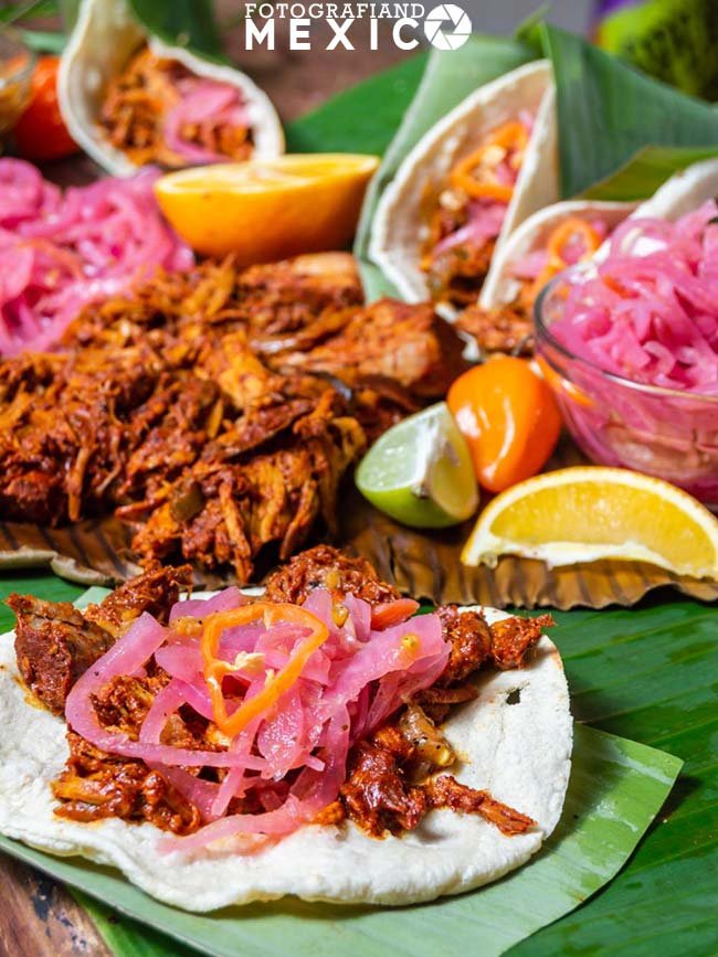 Carne de cochinita pibil servida sobre hojas de platano con cebolla morada curtida rodajas de naranja agria y chiles habaneros.