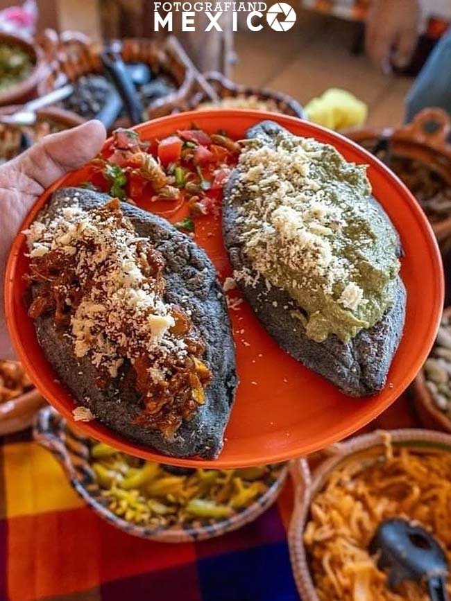 Dos tlacoyos de masa azul con carne deshebrada salsa verde queso rallado y nopales picados servidos en un plato naranja.