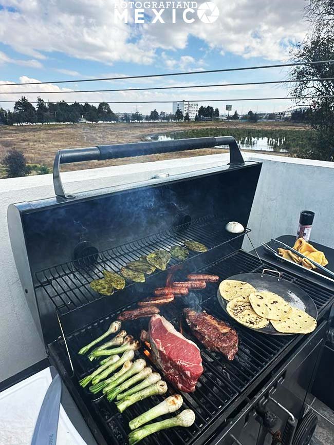 Cortes de carne asada con cebollitas nopales y tortillas calentandose sobre las rejillas de una parrilla abierta al aire libre.