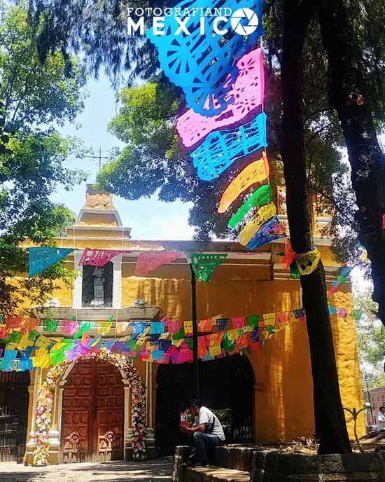 Fachada amarilla de la Capilla de Santa Catarina en Coyoacán decorada con papel picado de colores y flores en la entrada.